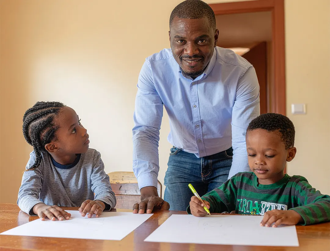 A father helping two children draw at a table at home